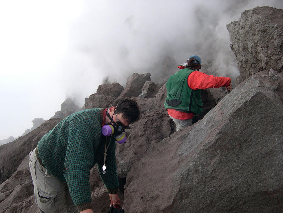 Two people, one in a gas mask and the other facing away, climb on a dark rocky mountain as smoke blows behind them.