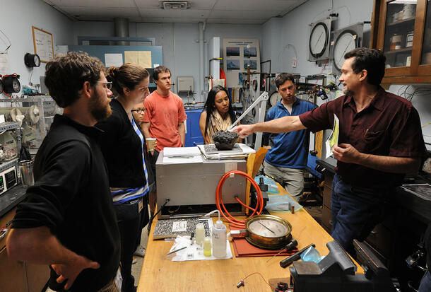 Six students gather around a table as curator Jim Webster speaks to them and points at rock specimen on the table.