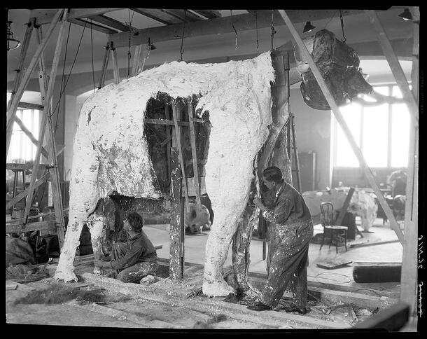 Two men in coveralls work on the legs of a wood and metal frame shaped like an elephant partially covered in white plaster.