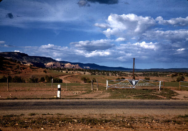 Gate with a bull image at the center and lettering reading Ghost Ranch in open landscape with hills in the background.