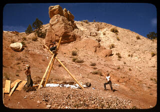 To people stand to the side as a person sits at the center of three wooden beams creating a tripod shape against rocky landscape.