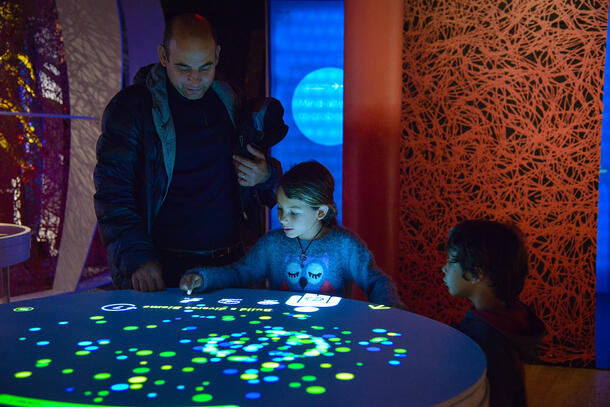 Two children and an adult look at and interact with a digital Museum exhibit about microbes.