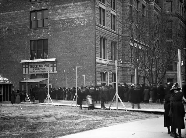 Historical image of long line of people waiting to enter building with sign reading: International Tuberculosis Exhibition. 