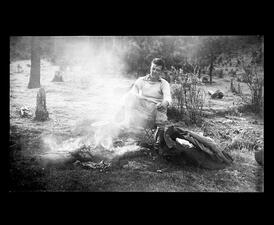 Alfred Kinsey sitting in foldable chair and warming one bare foot by campfire, among grass and trees.