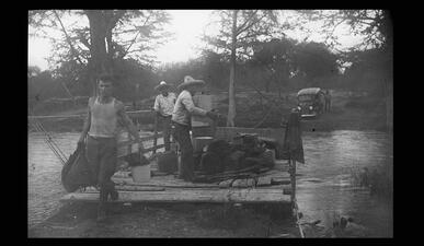 A man gets off of a river raft as two men in large hats remain on the raft, with one carrying cargo. 