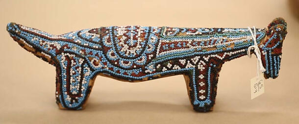 Stylized, colorfully beaded deer figurine with a long body and paper label on neck.