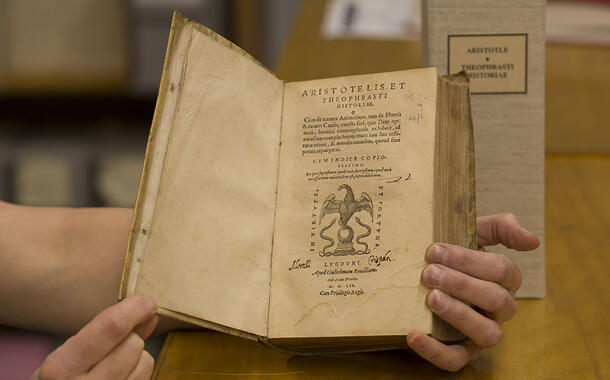 Hands hold open an old book to title page reading Aristotelis Et Theophrasti Historiae, plus additional text and illustration of bird and snakes.