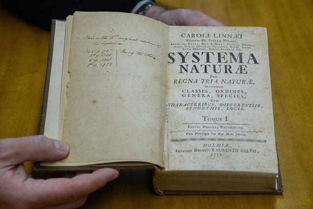 Hands holding open an old book to the title page reading Caroli Linnae, Systema Naturae and additional text.