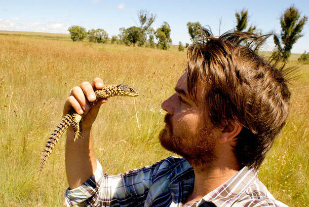 Person in a field holding a small lizard in his right hand close up to their face.