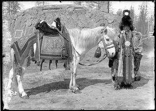 Woman in large fur headpiece, fur robe and large neck jewelry stands next to a white horse with an intricately decorated saddle. .