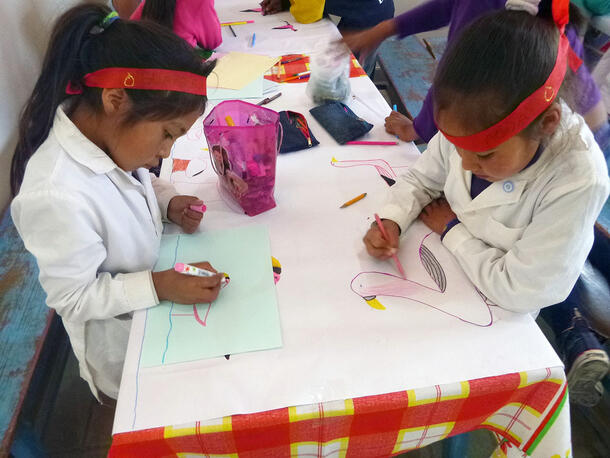 Two schoolchildren seated at a table draw pink birds with markers and colored pencil.
