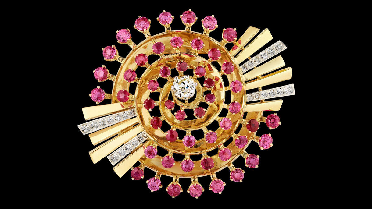 A bejeweled spiral clip studded with colorful rubies, a central diamond and ray-like pieces jutting out from two sides.