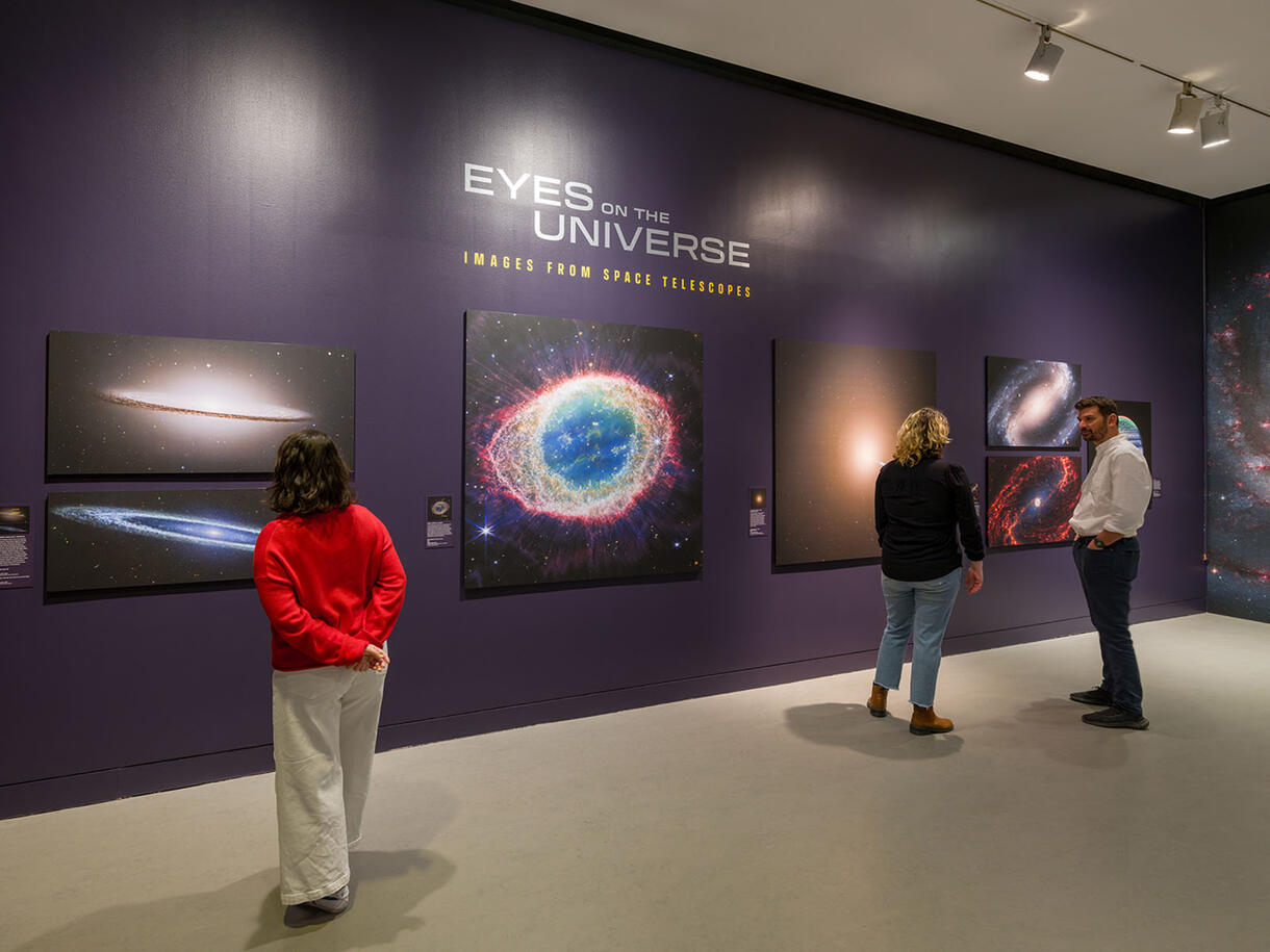 Three people stand in a gallery looking at large photos of space, with text "Eyes on the Universe: Images from Space Telescopes" printed on the wall.