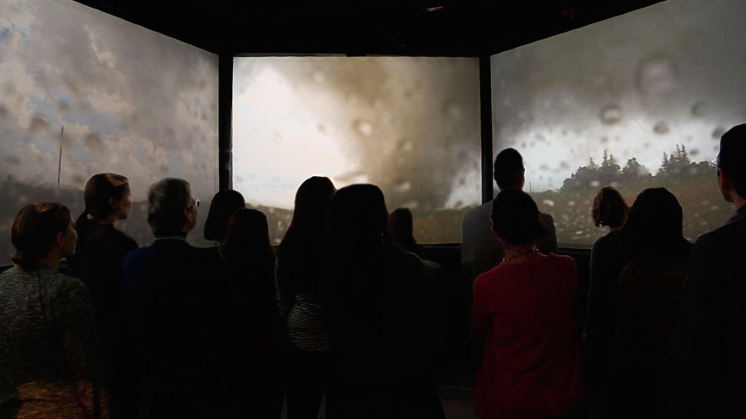 About thirteen visitors in silhouette face three screens playing video of a storm.