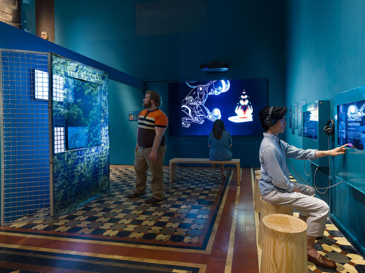 Three Museum visitors interact with video and audio components of the exhibitShaping the Future Through Tradition in the Northwest Coast Hall.