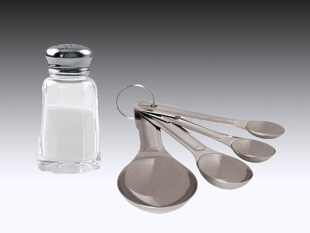 A salt shaker on a surface next to four measuring spoons.