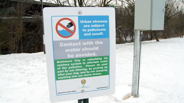 Metal sign posted on snowy ground, stating urban streams are subject to pollutants and runoff, urging against littering, dumping in storm drains, and contact with the water.