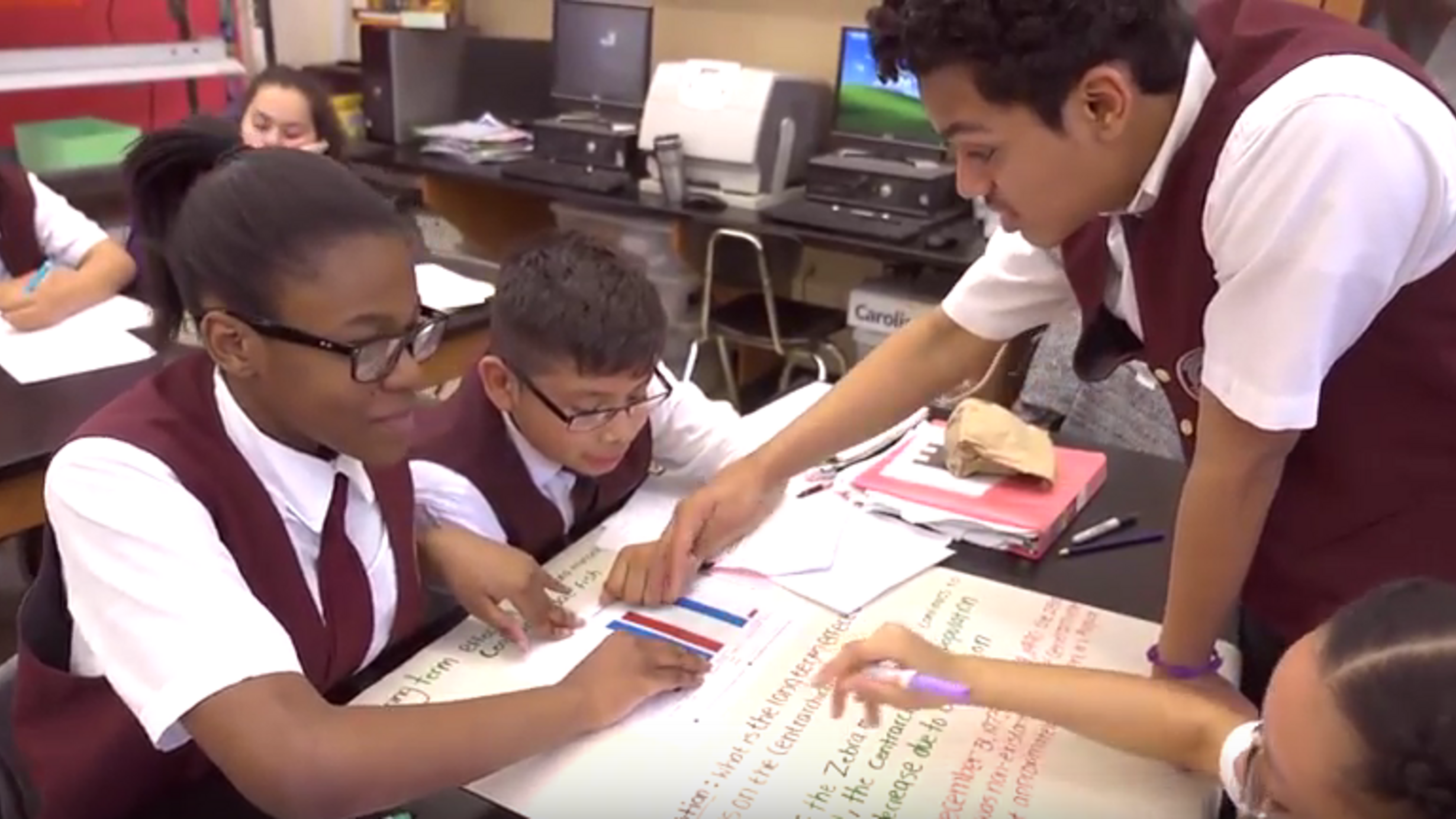 Four students in uniforms lean over a table as they work on a poster featuring text and a bar graph together.