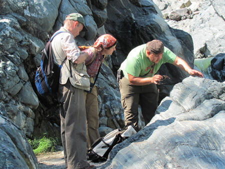 Two men and one woman, Kennet Flores, George E. Harlow, and Jinny Sisson outdoors examining a large rock formation. Flores discusses veins and layering in the formation.