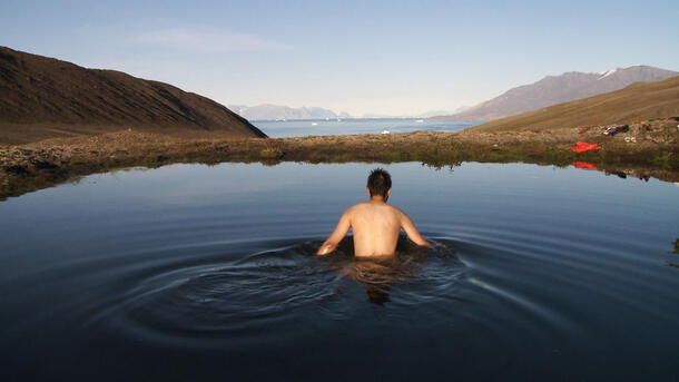 A young man with short dark hair, his bare back to the camera, wades waist-deep in a remote lake in Northern Greenland, surrounded by low hills, rolling tundra, and big clear sky.