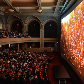 Movie theater pictured from the side with two levels of a full, seated audience watching fireworks display on the screen.