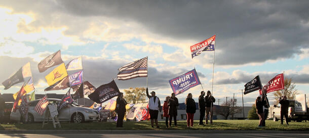 About ten people stand on the side of a road, some holding Trump 2020 campaign flags and one holding the "Thin Blue Line" American flag.