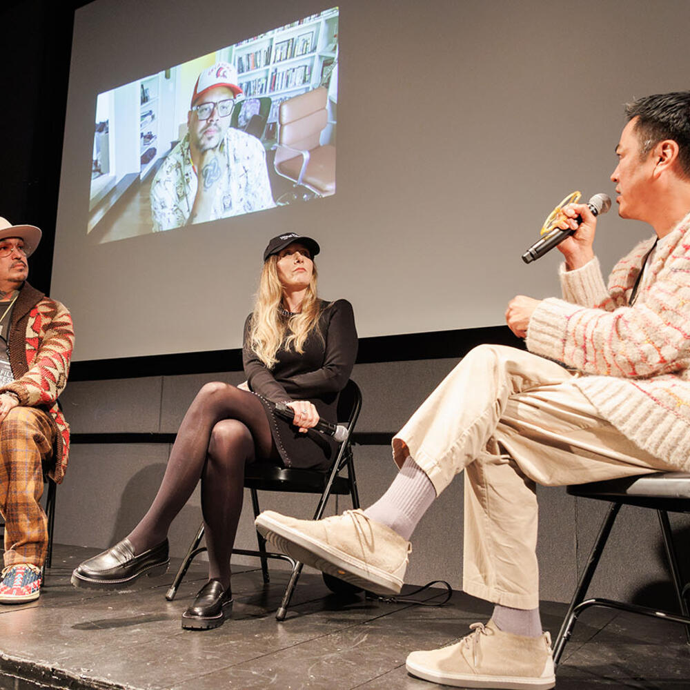 Billy Luther (right) speaks into a microphone to Erica Tremblay and Steven Paul Judd (seated on stage) and Sterlin Harjo (on screen behind them).