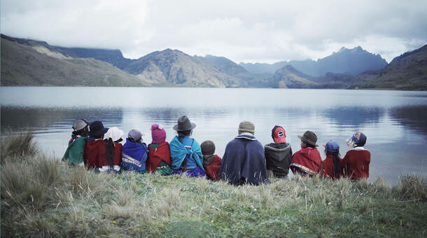 12 people, two adults and ten children, pictured from behind as they sit on grass beside a lake with mountains in the background.