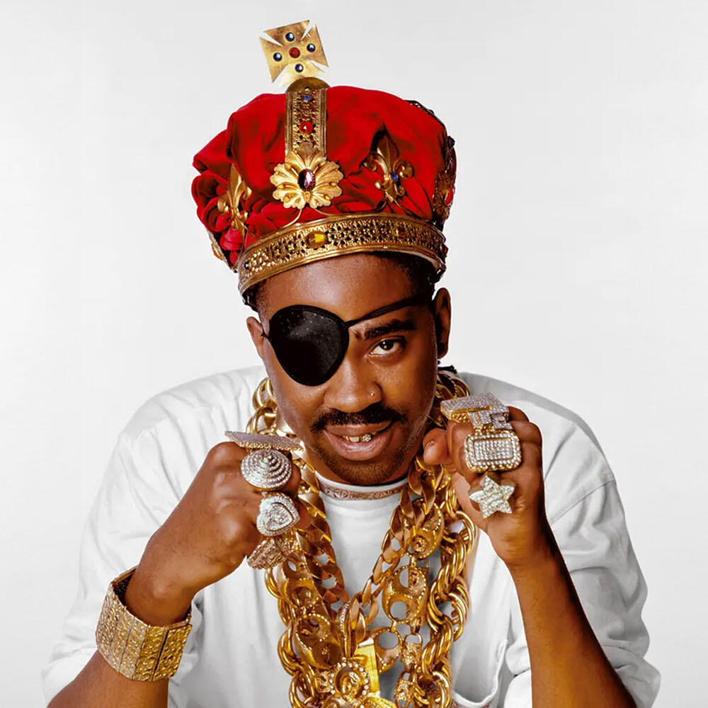 Slick Rick wearing an eyepatch over his right eye, a large crown, heavy gold necklaces, and over 7 large rings.