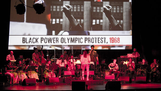 17 people on a stage, playing various instruments, below a screen with an image of two raised fists and text "Black Power Olympic Protest, 1968." 