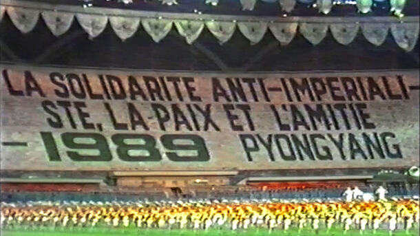 A stadium crowd display a tifo reading "La Solidarite Anti-Imperialiste, La Paix Et L'amitie - 1989 Pyongyang" as a large group perform on the stadium grass.