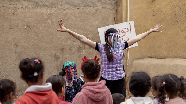 A young theater performer stands in front of a crowd of children with their arms out and a fringed headpiece covering their face.