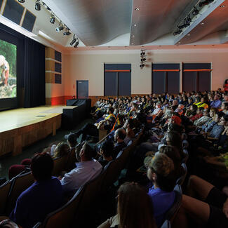 A seated audience in Kaufmann Theater watch a screen displaying an image of four people in conical straw hats working outside.