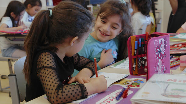 Two schoolchildren sit at a desk, one smiling and the other turned away from the camera using a colored pencil on a workbook.
