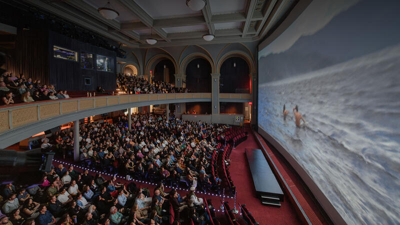 A seated audience in the LeFrak Theater watch the screen which shows two people waist deep in water with mountains in the background.