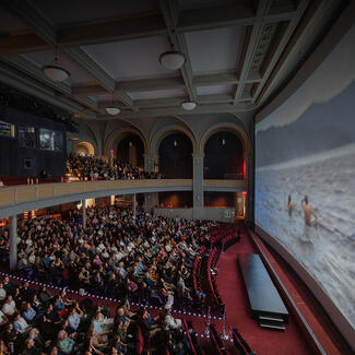 A seated audience in the LeFrak Theater watch the screen which shows two people waist deep in water with mountains in the background.