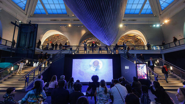 A crowd of visitors sit in the Milstein Hall of Ocean Life beneath the whale watching an animated short on a screen in an alcove between staircases.