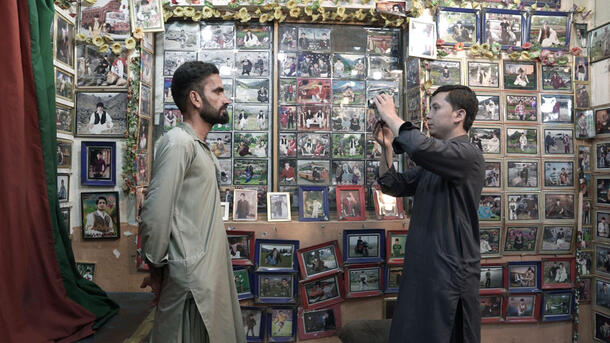 A side-view of a photographer holding a camera up to take a photo of a client with many framed photos decorating the wall behind them.