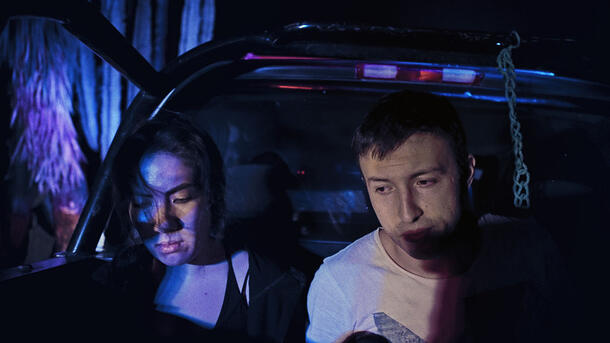 Two people sit in the open trunk of a car, their faces illuminated by an artificial light at night. 