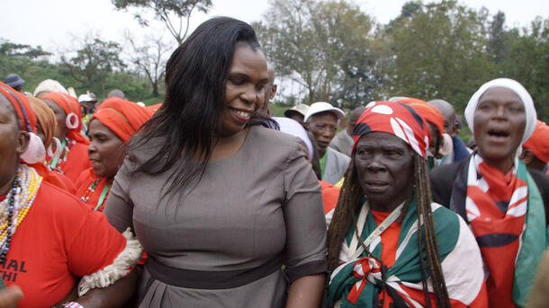 Wanjugu Kimathi walks arm in arm with two people, one an older person wearing a scarf on their head, ahead of a few more rows of people.
