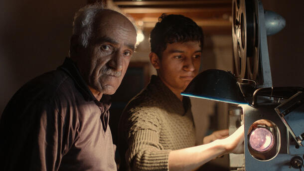An older man, Samid, and the younger Ayaz stand in front of a film projector, with Samid looking directly at the camera.