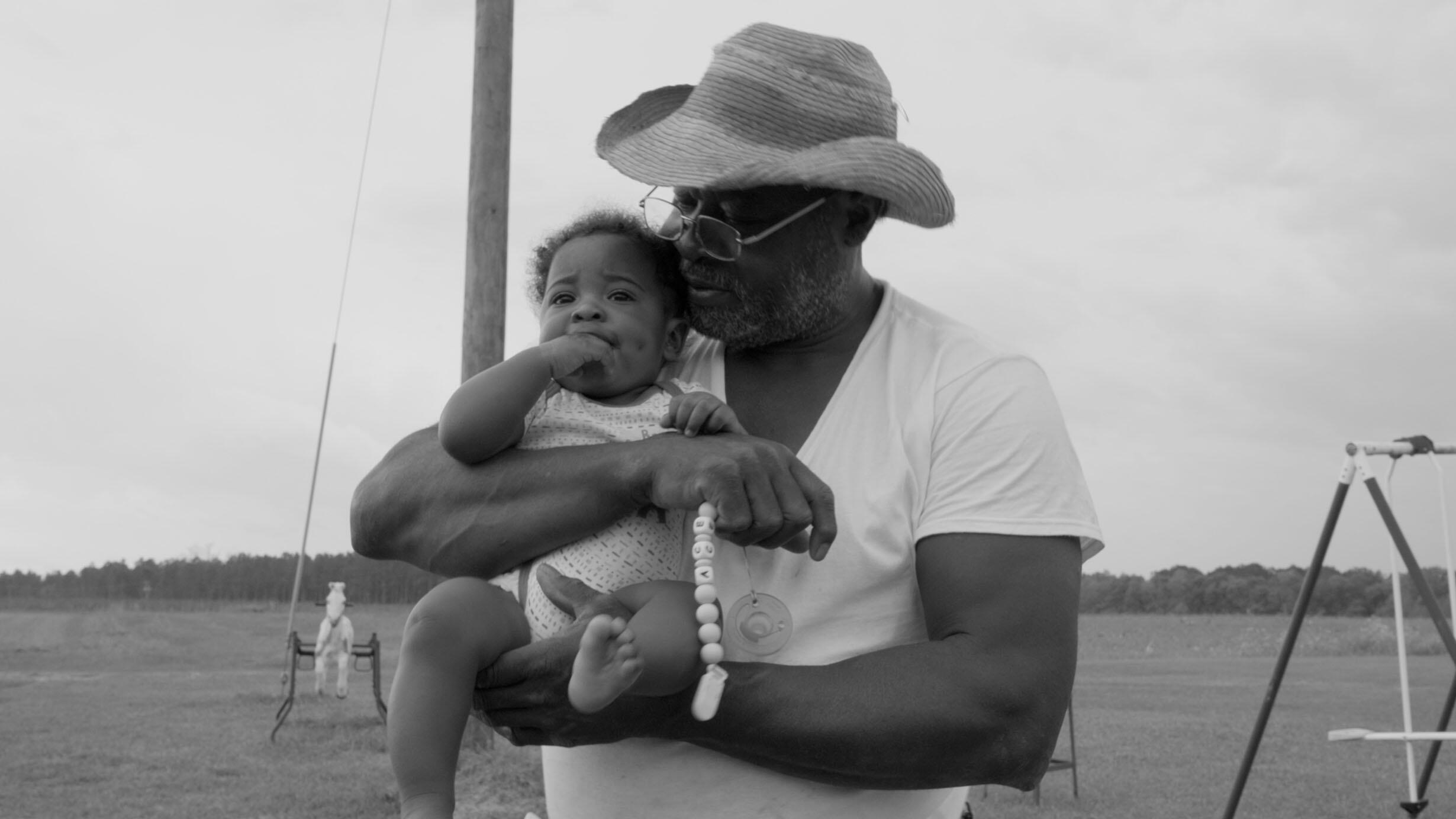 Farmer Willie Head Jr., wearing a wide-brimmed straw hat, holds a baby and a pacifier.