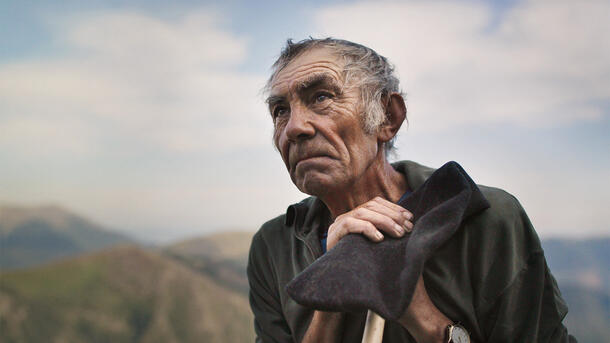 Close-up on Yves Raspaud, an aging shepherd, gazing into the distance and leaning on a walking stick with mountains visible in the background.
