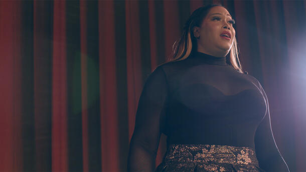 An opera singer wearing a turtleneck and patterned bottoms stands on a stage in front of a curtain with light shining in their face.