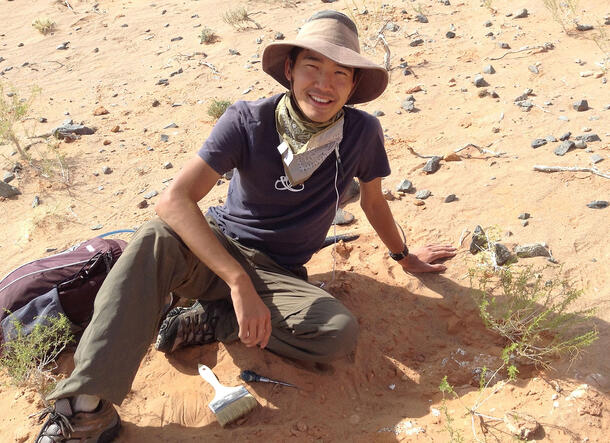 Akino wears a hat and bandana and sits in the sand beside his tools (a pick and a brush).