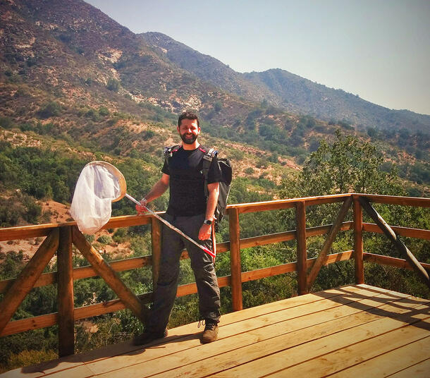 Santos, standing on a wooden deck in a mountainous area, holds a net and wears a backpack.
