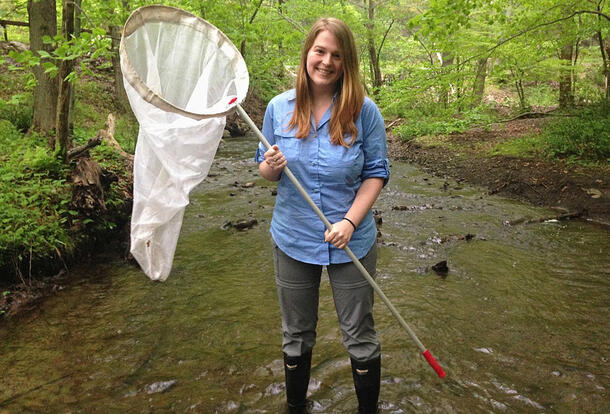 Oliver stands in a stream, wearing rubber boots and carrying a net.