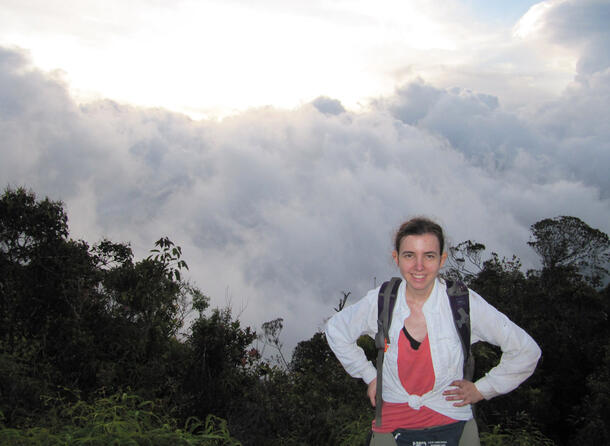 Loria stands in front of trees with light breaking through the clouds in the background.