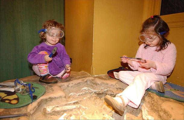 Two children with googles and tools perch on a sand pit that holds "fossils" waiting to be discovered.