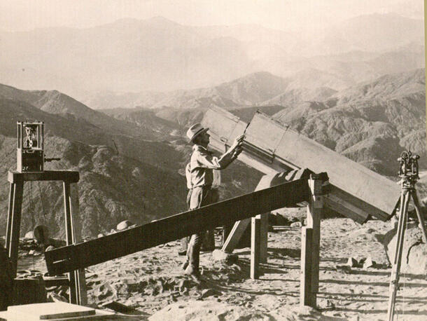 Person stands at the of a mountain looking into a very large telescope.